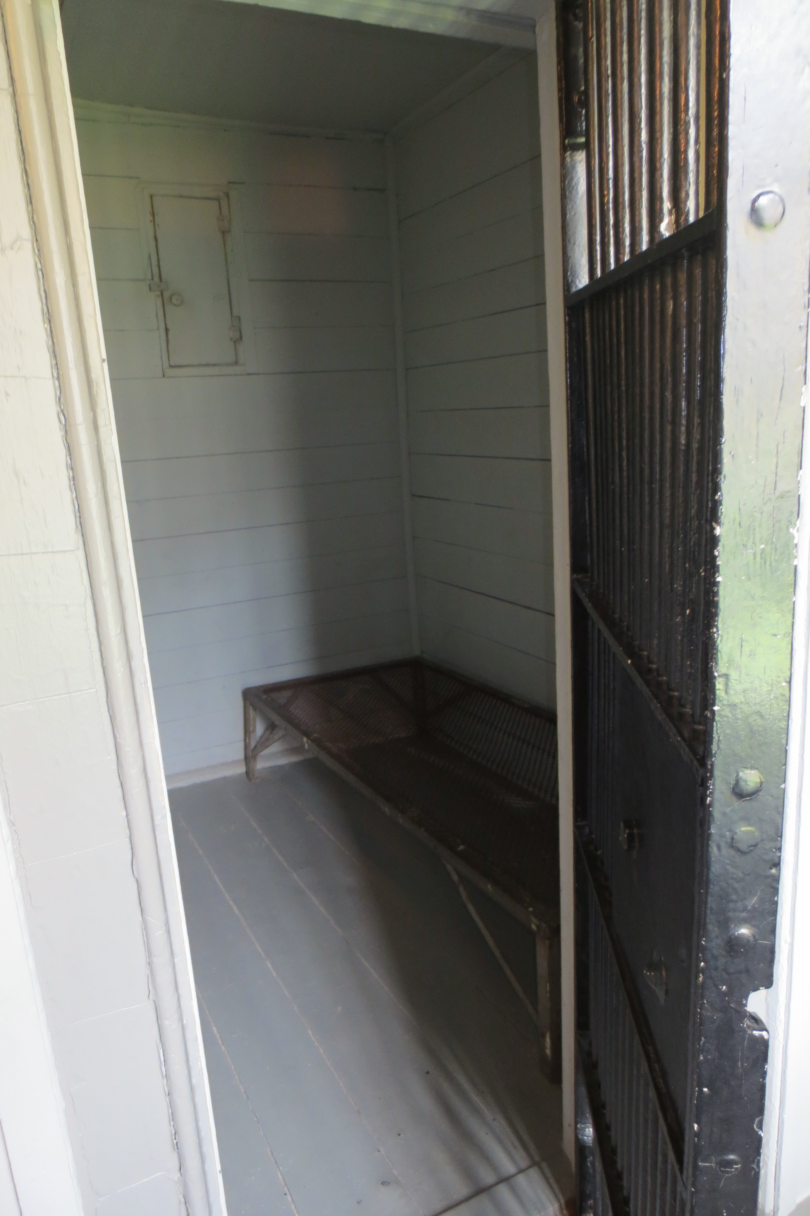 Photograph of a jail cell, barred door ajar. Metal bead inside. Small rectangular window closed.