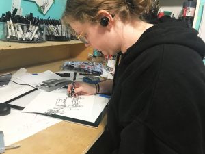 Image of a woman in a black hoodie hunched over a desk drawing with ink pen.