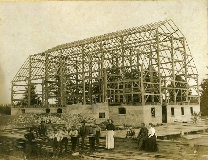 Black and white photograph of a barn raising. Bank Barn. Frame and foundation in place. Men and women standing in front.